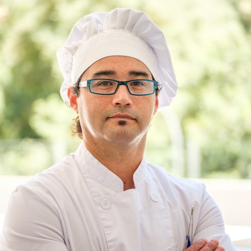 Professional male chef wearing glasses and toque blanche, standing confidently with crossed arms.